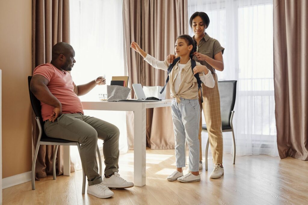 A parent helps a child put on a school backpack at home while another parent sits at the table during the morning routine