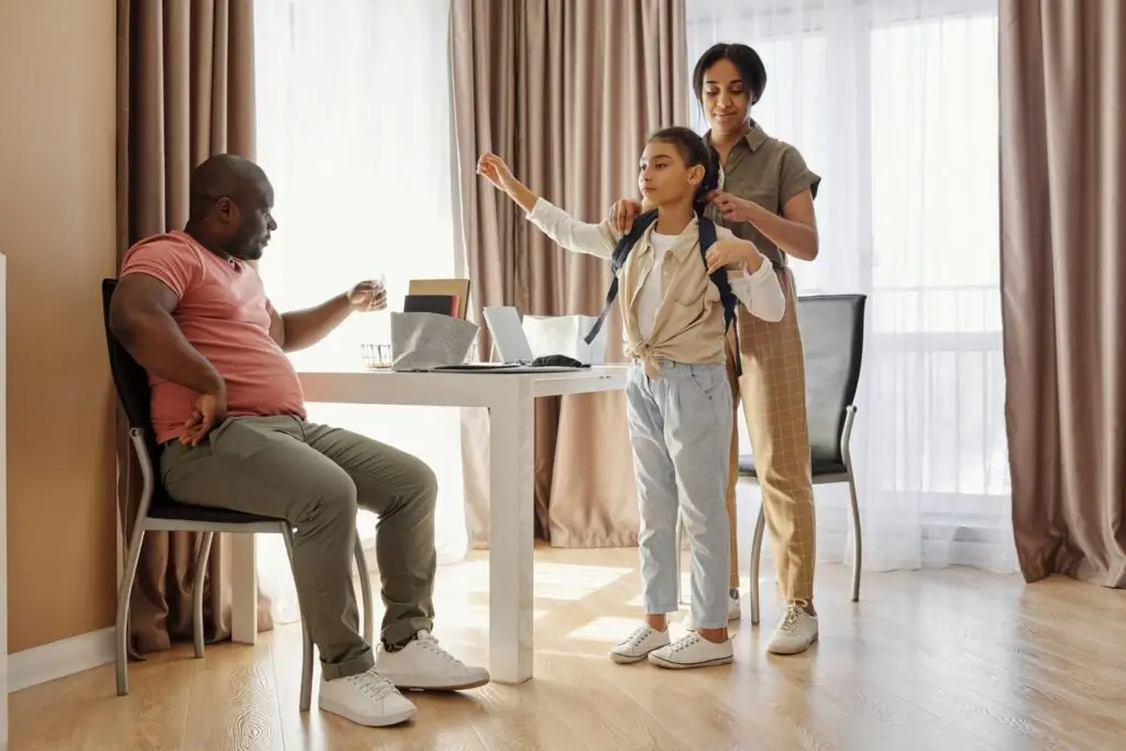 A parent helps a child put on a school backpack at home while another parent sits at the table during the morning routine