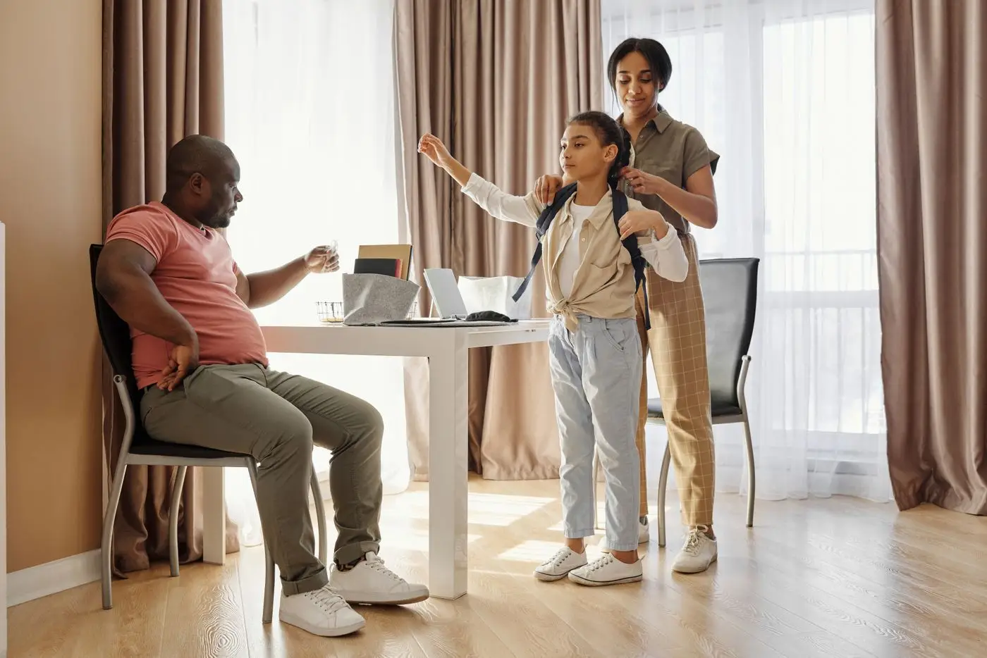 A parent helps a child put on a school backpack at home while another parent sits at the table during the morning routine