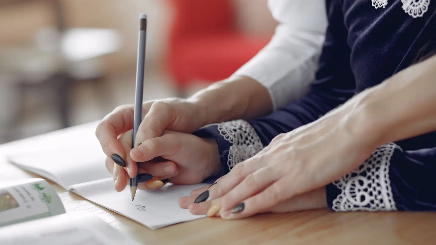 An adult's hand guides a child's hand as they write in a lined notebook at a wooden table.