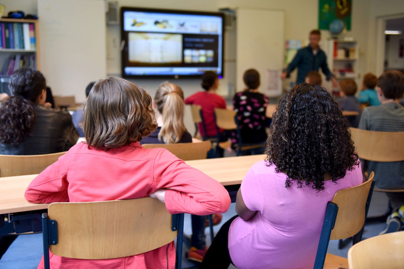 Primary school students seated at desks watching a teacher present a lesson on a screen in a bright international school classroom