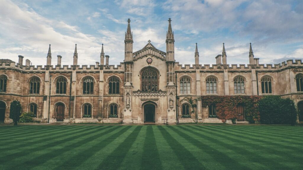 A traditional English collegiate building in pale stone behind a striped green lawn under a blue sky.