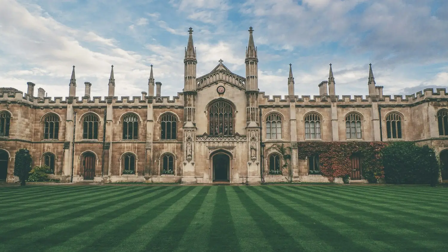 A traditional English collegiate building in pale stone behind a striped green lawn under a blue sky.