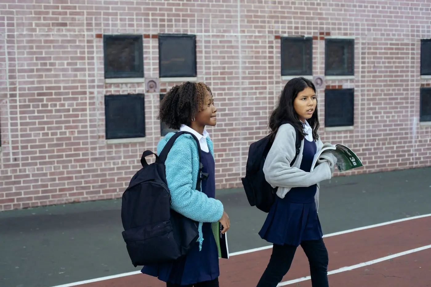 Two schoolgirls in uniform with backpacks walking past a red-brick school building on their way into class