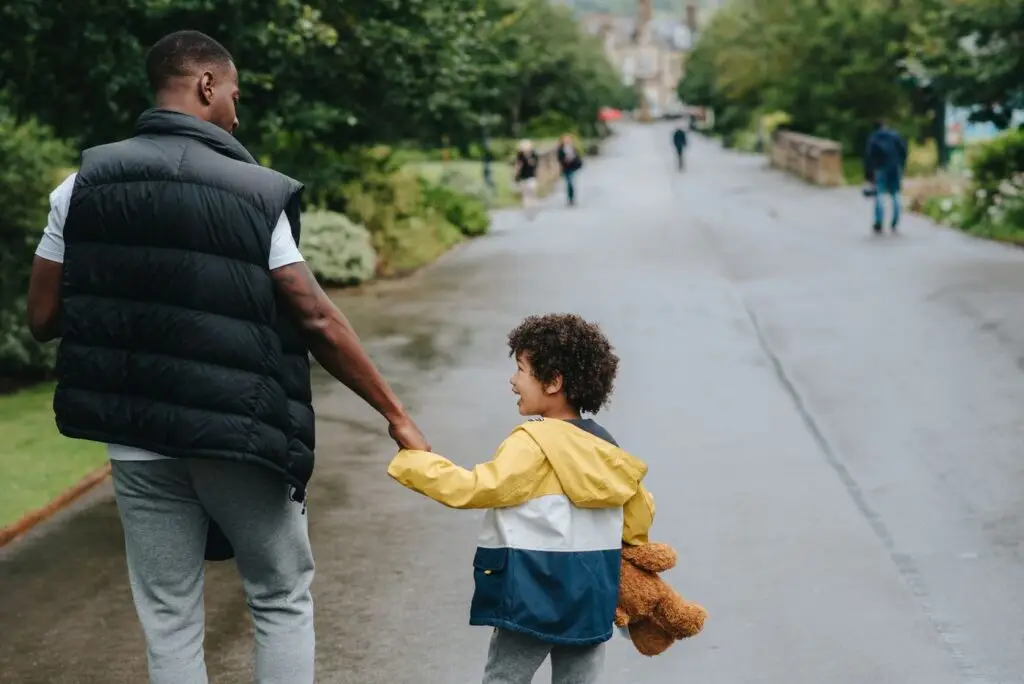 A father and young son walking hand in hand along a wet tree-lined path on an overcast day, the child carrying a teddy bear