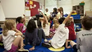 Young children sit on a classroom carpet with their hands raised while a teacher reads to them; French immersion in Toronto begins at this age, in Junior Kindergarten.